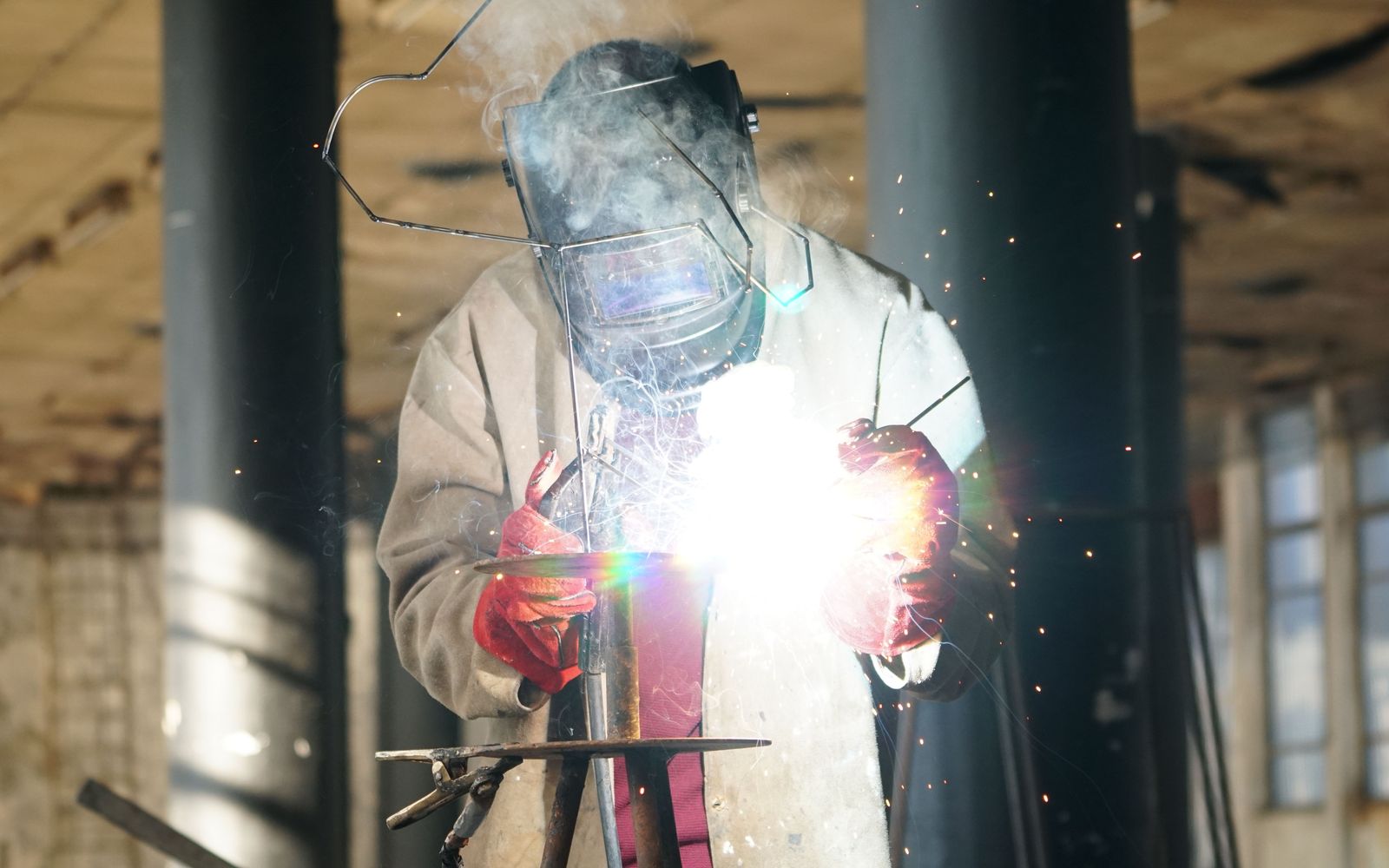 A welder in a full helmet and red gloves welding steel, sparks flying in a daylit hall